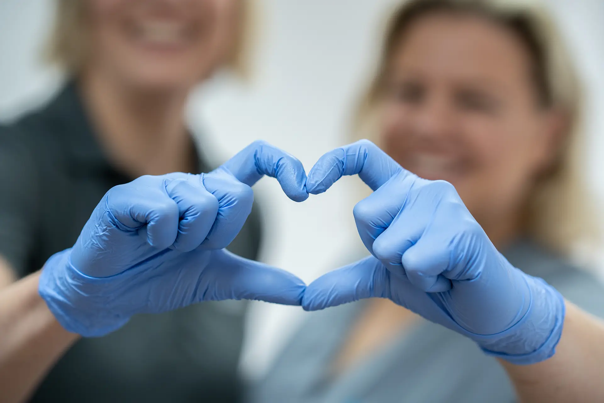 Zwei Mitarbeiterinnen mit blauen Handschuhen formen mit den Händen ein Herz